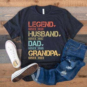 May include: A black t-shirt with a vintage-style graphic that reads "LEGEND SINCE 1959, HUSBAND SINCE 1987, DAD SINCE 1989, GRANDPA SINCE 2022". The graphic is in various colors, including blue, green, and yellow.