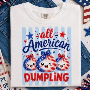 Op de afbeelding: Wit T-shirt met een patriottisch ontwerp met de tekst "all American DUMPLING". De afbeelding bevat drie dumpling-personages met rood, wit en blauw gestreepte patronen en strikken, samen met sterren. Het shirt wordt getoond op een denim achtergrond.