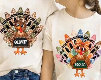 May include: Two cream-colored t-shirts featuring turkey graphics made of crayons. The left turkey wears a cap and holds a sign reading OLIVER. The right turkey wears a bow and holds a sign reading SOPHIA. Both turkeys have colorful, patterned crayon tail feathers.