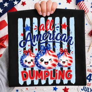 Op de afbeelding: Zwart T-shirt met een patriottisch ontwerp. Het shirt heeft de tekst "All American Dumpling" in rood en blauw, met drie dumpling-personages versierd met de Amerikaanse vlag. Blauwe en witte strepen en sterren maken het ontwerp compleet.