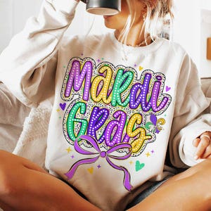 May include: A white sweatshirt with a colourful "Mardi Gras" graphic. The graphic features purple, green and gold lettering with white polka dots. The letters are outlined in black and have a light yellow glow.