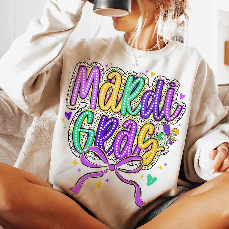 May include: A white sweatshirt with a colourful "Mardi Gras" graphic. The graphic features purple, green and gold lettering with white polka dots. The letters are outlined in black and have a light yellow glow.