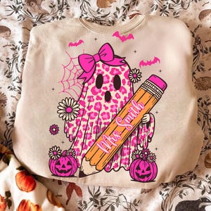 May include: Beige sweatshirt with a pink leopard print ghost design, holding a pencil with "Mrs. Smith" written on it. The ghost has a pink bow, and the design includes bats, spiderwebs, daisies, and jack-o'-lanterns. A fun Halloween or teacher-themed design.