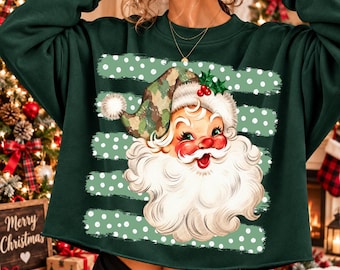 May include: A dark green long-sleeve sweatshirt featuring a graphic of a Santa Claus face with a camouflage-patterned hat. The design includes horizontal light green stripes with white polka dots behind the Santa illustration, which has a white beard.