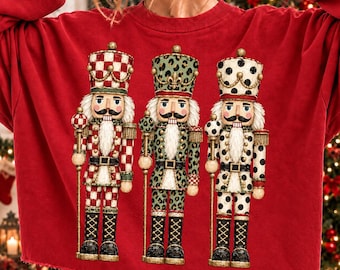 May include: A red long-sleeve sweatshirt featuring a graphic print of three nutcrackers. The left nutcracker has a red and white checkered pattern, the middle has a green leopard print, and the right has a white and black polka dot pattern.