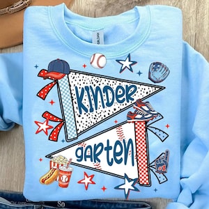 May include: A light blue crewneck sweatshirt featuring a graphic print of two pennants with the text "KINDER" and "GARTEN". The design includes baseball-themed illustrations such as a baseball, a cap, a glove, cleats, a hot dog, and a drink.