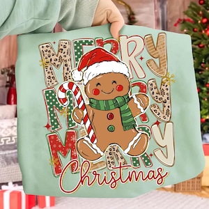 Può includere: Una borsa tote verde chiaro con un omino di pan di zenzero con cappello e sciarpa di Babbo Natale, che tiene un bastoncino di zucchero. Le parole "Merry Christmas" sono stampate in un design festivo con accenti animalier.