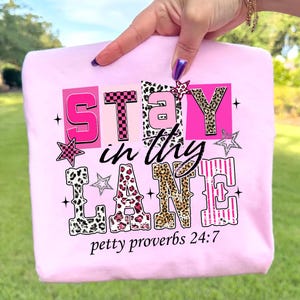 May include: A light pink sweatshirt with the text "STAY in thy LANE" in a mix of fonts and patterns, including leopard print and stripes. The phrase "petty proverbs 24:7" is printed below the main text. The design includes star accents.