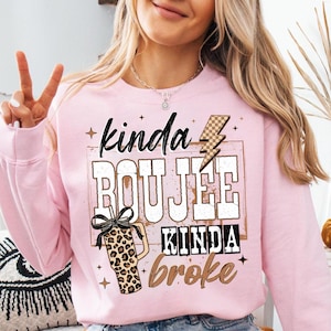 May include: A light pink sweatshirt with the text "kinda BOUJEE kinda broke" in a mix of fonts and colors. A leopard print tumbler with a black bow is on the left. A lightning bolt graphic is above the word "BOUJEE".