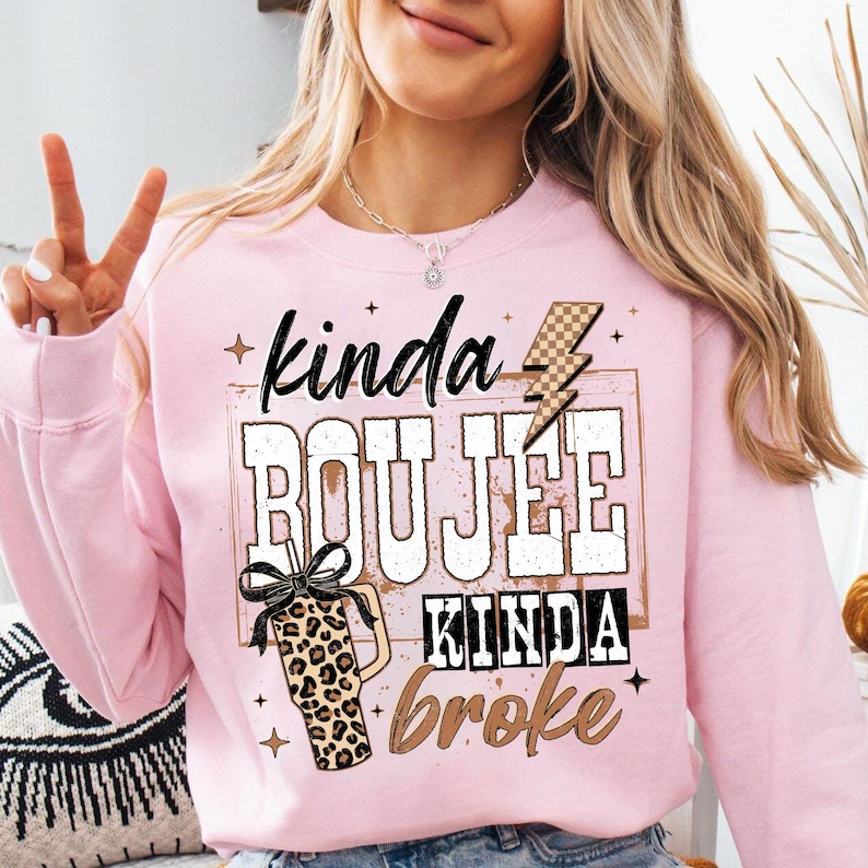 May include: A light pink sweatshirt with the text "kinda BOUJEE kinda broke" in a mix of fonts and colors. A leopard print tumbler with a black bow is on the left. A lightning bolt graphic is above the word "BOUJEE".