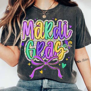 May include: A black t-shirt featuring a purple, green and yellow "Mardi Gras" design with a purple bow and glitter accents.