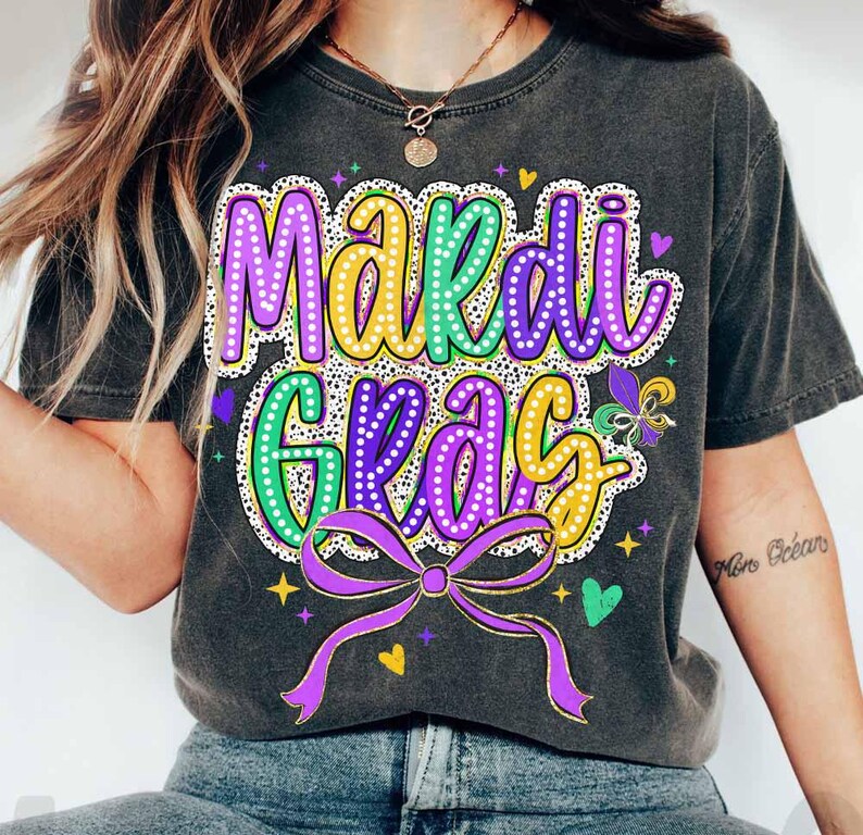 May include: A black t-shirt featuring a purple, green and yellow "Mardi Gras" design with a purple bow and glitter accents.