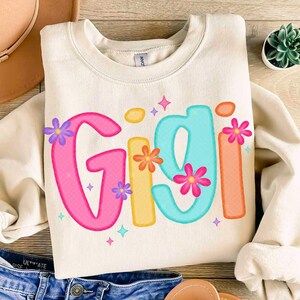 Peut inclure: Un sweat-shirt blanc avec le nom "Gigi" &eacute;crit en lettres color&eacute;es et soulign&eacute;es. Les lettres sont d&eacute;cor&eacute;es de petites fleurs color&eacute;es. Le sweat-shirt est de couleur cr&egrave;me clair et a un col rond.