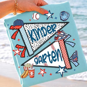 May include: A light blue t-shirt featuring two pennant flags with the text "KINDER" and "GARTEN" in blue script. The design includes baseball-themed graphics like a baseball, glove, cleats, a hot dog, popcorn, and stars in red and blue.