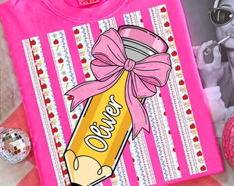 May include: A bright pink t-shirt featuring a graphic of a yellow pencil with a large pink bow tied around it. The pencil has the name Oliver printed on it. The background of the graphic consists of vertical stripes with small red apple icons.
