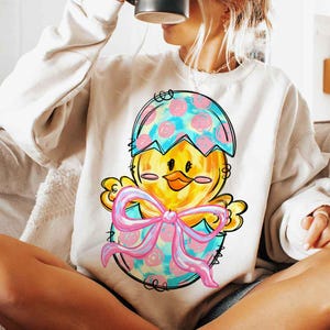 May include: A white sweatshirt with a colorful cartoon chick hatching from an egg. The chick is yellow with a pink bow and the egg is blue, pink, and white with a polka dot pattern.