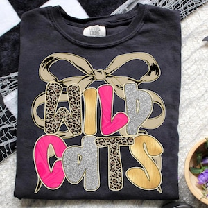 May include: Dark gray t-shirt with the text "WILD CATS" in a layered design. The letters feature leopard print, pink, silver glitter, and gold colors, with a tan bow graphic behind the text. The shirt is a Comfort Colors brand.