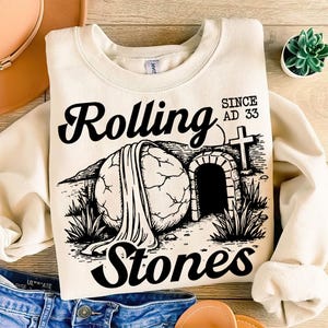 May include: A beige sweatshirt featuring a black graphic of a stone rolling away from a tomb. The graphic includes the text "Rolling Stones" and "Since AD 33".