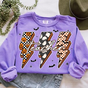 May include: A light purple sweatshirt with a Halloween-themed design. The design features three lightning bolt shapes with different patterns: jack-o'-lanterns, ghosts, and bats. The word "Teacher" is written across the bolts.
