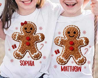 May include: White t-shirts featuring a gingerbread man design. One shirt has the name "Sophia" in red, and the other has "Matthew". The gingerbread men are brown with white icing, red buttons, and a red bow. Christmas-themed.