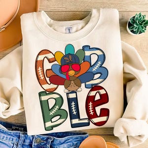 May include: A white sweatshirt with a Thanksgiving-themed design. The design features a cartoon turkey wearing sunglasses and a football helmet, with the word "Gobble" written in colorful letters with football patterns.