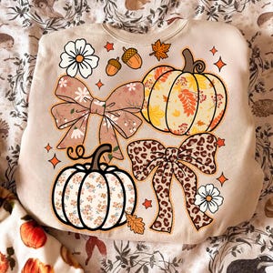 May include: Beige sweatshirt with fall-themed graphics. Features a pumpkin, bows, flowers, acorns, and leaves. The pumpkin and bow designs have floral and leopard prints. Orange stars and a white flower accent the design.