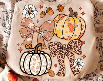 May include: Beige sweatshirt with fall-themed graphics. Features a pumpkin, bows, flowers, acorns, and leaves. The pumpkin and bow designs have floral and leopard prints. Orange stars and a white flower accent the design.