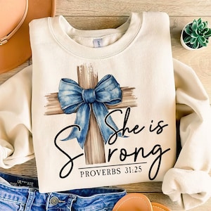 May include: A cream-colored sweatshirt with a brown wooden cross with a blue bow on it. The text "She is Strong" and "Proverbs 31:25" are printed below the cross.