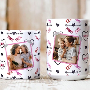 Op de afbeelding: Twee witte keramische mokken met een zwart en roze ontwerp. De mokken hebben een foto van een moeder en kind, harten, pijlen en de tekst "Best Mom Ever".