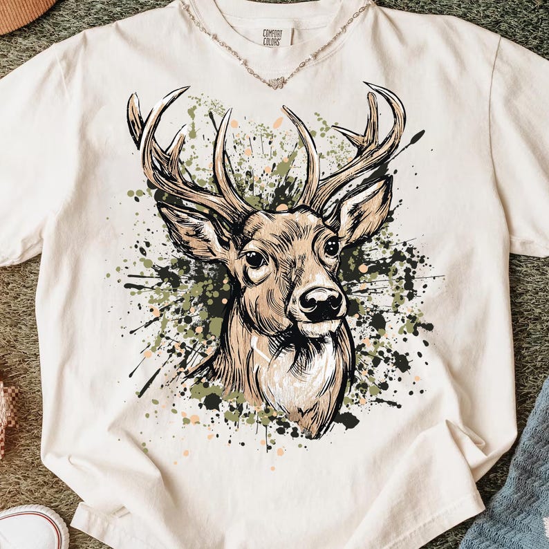 May include: Cream-colored t-shirt featuring a detailed illustration of a deer's head with large antlers. The design includes a splatter effect in shades of green, black, and brown. The shirt has a classic crew neck and short sleeves. The brand name "COMFORT COLORS" is printed on the collar.