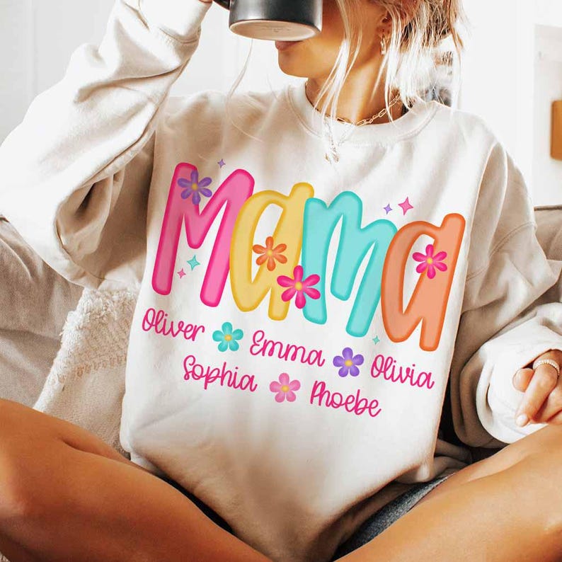 Peut inclure: Sweat-shirt blanc avec le mot "Mama" en lettres color&eacute;es avec des fleurs et des paillettes. Sous le mot "Mama" se trouvent les noms Oliver, Emma, Sophia et Phoebe.