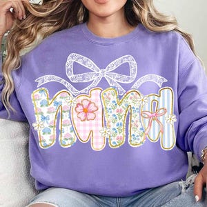 K&ouml;nnte beinhalten: Lilafarbener Pullover mit einem floralen Design, das im Patchwork-Stil "Mama" sagt. Das Design enth&auml;lt eine wei&szlig;e Schleife aus Spitze und rosa, blaue und wei&szlig;e Blumen.