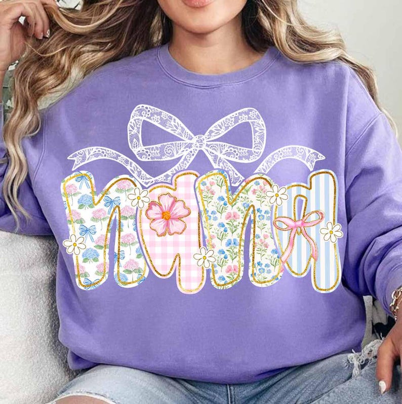 K&ouml;nnte beinhalten: Lilafarbener Pullover mit einem floralen Design, das im Patchwork-Stil "Mama" sagt. Das Design enth&auml;lt eine wei&szlig;e Schleife aus Spitze und rosa, blaue und wei&szlig;e Blumen.