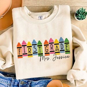 Op de afbeelding: Een cr&egrave;mekleurig sweatshirt met de woorden "First Grade" gespeld in regenboogkleurige kleurpotloden. De tekst "Mrs. Jessica" staat onder de kleurpotloden.
