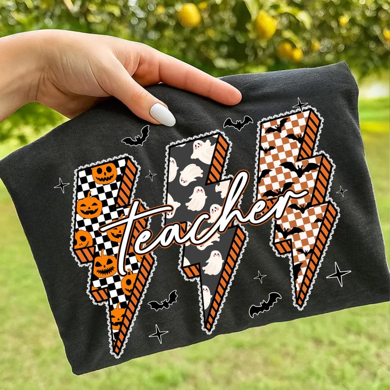 May include: Black t-shirt featuring a Halloween-themed design. The design includes three lightning bolt shapes with jack-o'-lanterns, ghosts, and bats. The word "Teacher" is written across the lightning bolts.