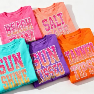 Op de afbeelding: Vijf kleurrijke t-shirts met zomerslogans. De shirts zijn roze, oranje, blauwgroen, paars en oranje. De tekst bevat "Beach Bum", "Salt Babe", "Sun Shine", "Sun Kissed" en "Tanned Tipsy".