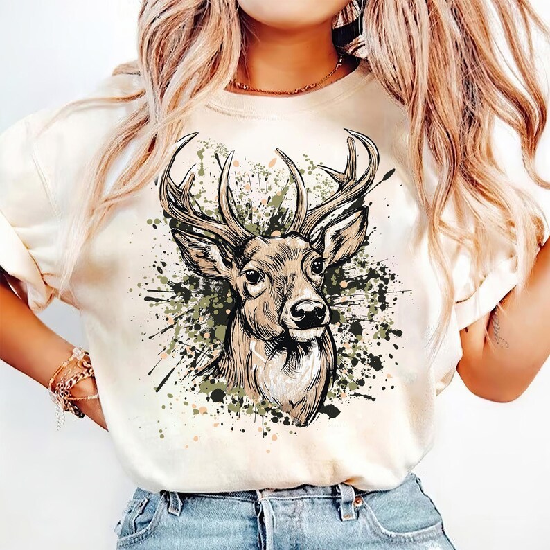 May include: Cream-colored t-shirt featuring a detailed illustration of a deer head with large antlers. The deer is depicted in shades of brown and tan, set against a backdrop of green and black paint splatters. A casual, nature-themed graphic tee.