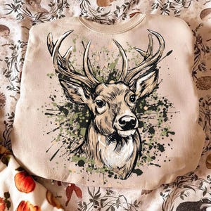 May include: A beige sweatshirt featuring a detailed illustration of a deer's head with large antlers. The deer is depicted in shades of brown and tan, with green and black paint splatters around it. The sweatshirt is made of a soft material.