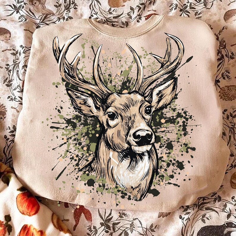 May include: A beige sweatshirt featuring a detailed illustration of a deer's head with large antlers. The deer is depicted in shades of brown and tan, with green and black paint splatters around it. The sweatshirt is made of a soft material.