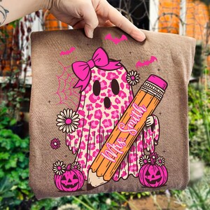 May include: A brown pillow with a Halloween-themed design. The design features a pink leopard print ghost wearing a bow, holding a pencil with the text "Mrs. Smith", and surrounded by pumpkins, bats, and flowers.