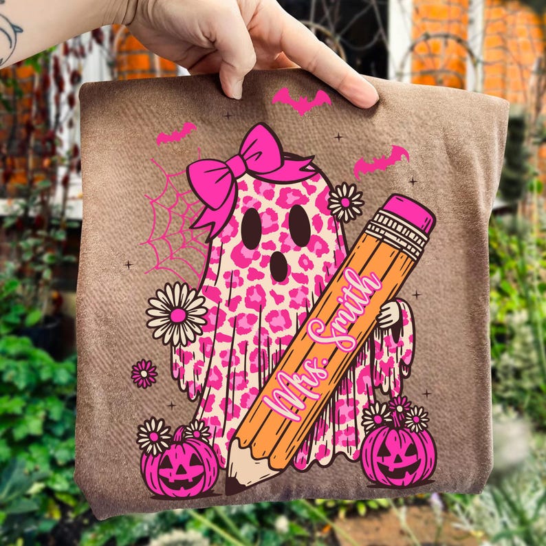 May include: A brown pillow with a Halloween-themed design. The design features a pink leopard print ghost wearing a bow, holding a pencil with the text "Mrs. Smith", and surrounded by pumpkins, bats, and flowers.