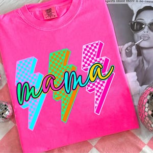 May include: Bright pink t-shirt with the word "mama" in a cursive font, overlaid on three lightning bolt graphics. The lightning bolts feature checkered patterns in shades of blue, green, and pink. The shirt has a crew neck and short sleeves.