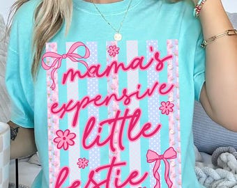 May include: A light turquoise t-shirt featuring a graphic print with vertical stripes in alternating shades of blue and white. The design includes the pink cursive text "mama's expensive little bestie," accented with pink bows and small flower icons.
