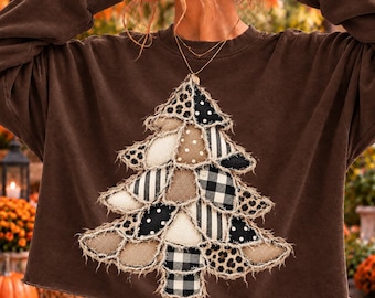 May include: A dark brown long-sleeve sweatshirt featuring a large, central Christmas tree applique. The tree is constructed from layered, frayed-edge fabric patches in various patterns, including black and white buffalo plaid, polka dots, and leopard print.