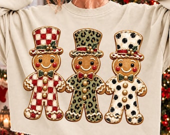 May include: A beige long-sleeve sweatshirt featuring a graphic of three gingerbread cookies wearing top hats. The first cookie has a red and white checkered pattern, the second has a green leopard print, and the third has black polka dots.