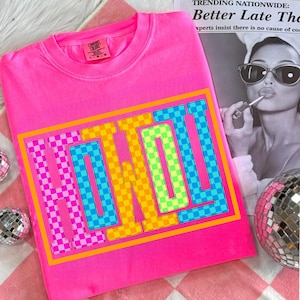 May include: Bright pink sweatshirt with the word "HOWWT" in a colorful checkered design. The letters are outlined in orange and framed by a yellow border. The sweatshirt is made by Comfort Colors.