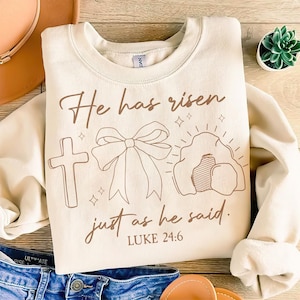 May include: A beige sweatshirt with a brown graphic design featuring a cross, a bow, and a tomb. The text "He has risen just as he said. LUKE 24:6" is printed on the sweatshirt.