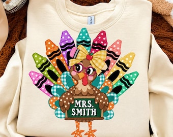 May include: A cream-colored crewneck sweatshirt featuring a graphic of a turkey with feathers made of colorful polka-dot crayons. The turkey wears pink glasses and a yellow plaid bow, holding a green chalkboard sign that reads MRS. SMITH.