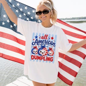 Op de afbeelding: Wit T-shirt met een afbeelding met de tekst "all American DUMPLING". De afbeelding toont drie dumpling-personages met patriottische rode, witte en blauwe ontwerpen. De persoon houdt een Amerikaanse vlag vast.