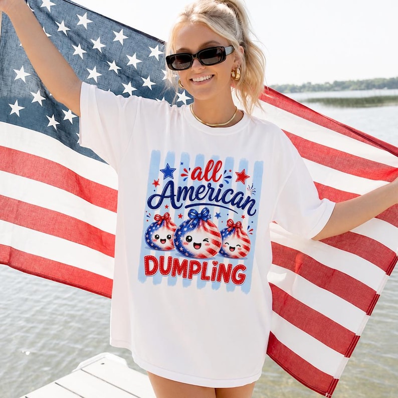 Op de afbeelding: Wit T-shirt met een afbeelding met de tekst "all American DUMPLING". De afbeelding toont drie dumpling-personages met patriottische rode, witte en blauwe ontwerpen. De persoon houdt een Amerikaanse vlag vast.